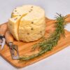 Tagliere di legno con sopra una forma circolare di formaggio fresco, con un ramoscello di ulivo accanto e due coltelli per formaggi.
